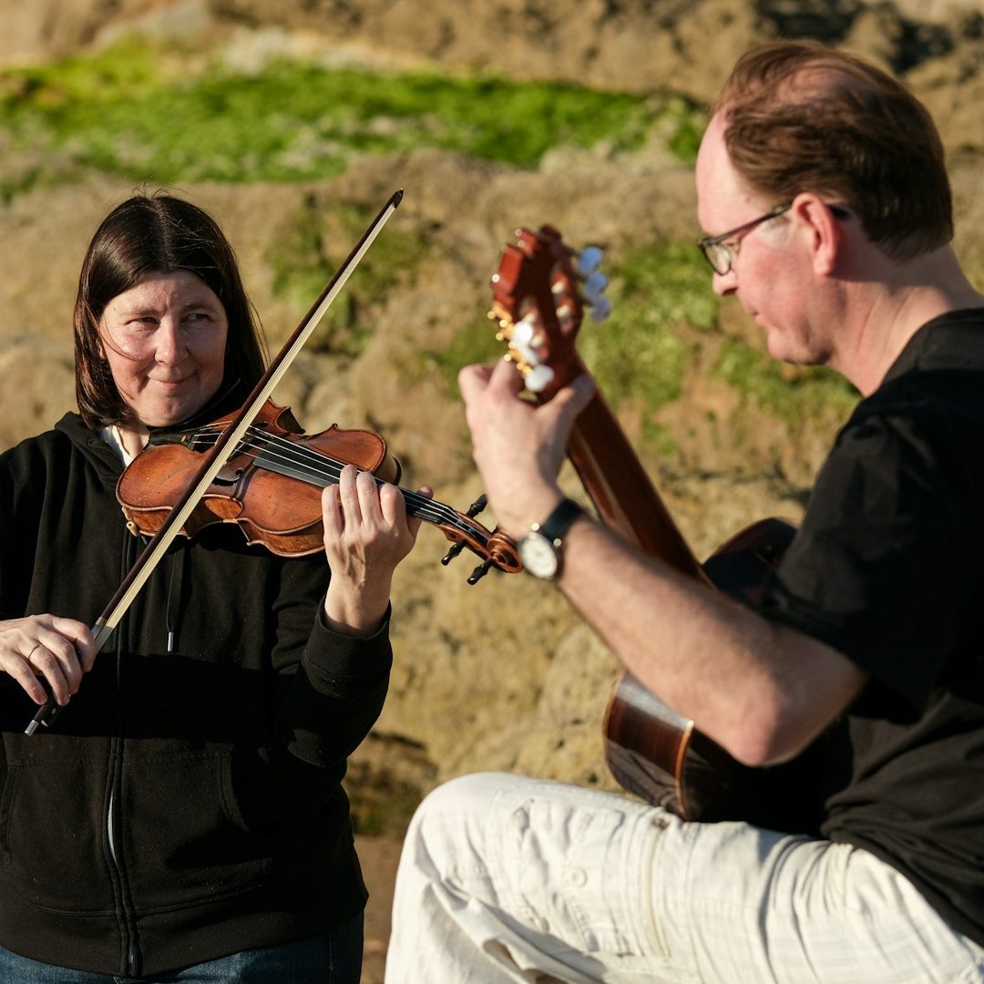 White woman wearing black hoodie and jeans plays violin and a man wearing a black shirt &white pants and glasses plays guitar outdoors.