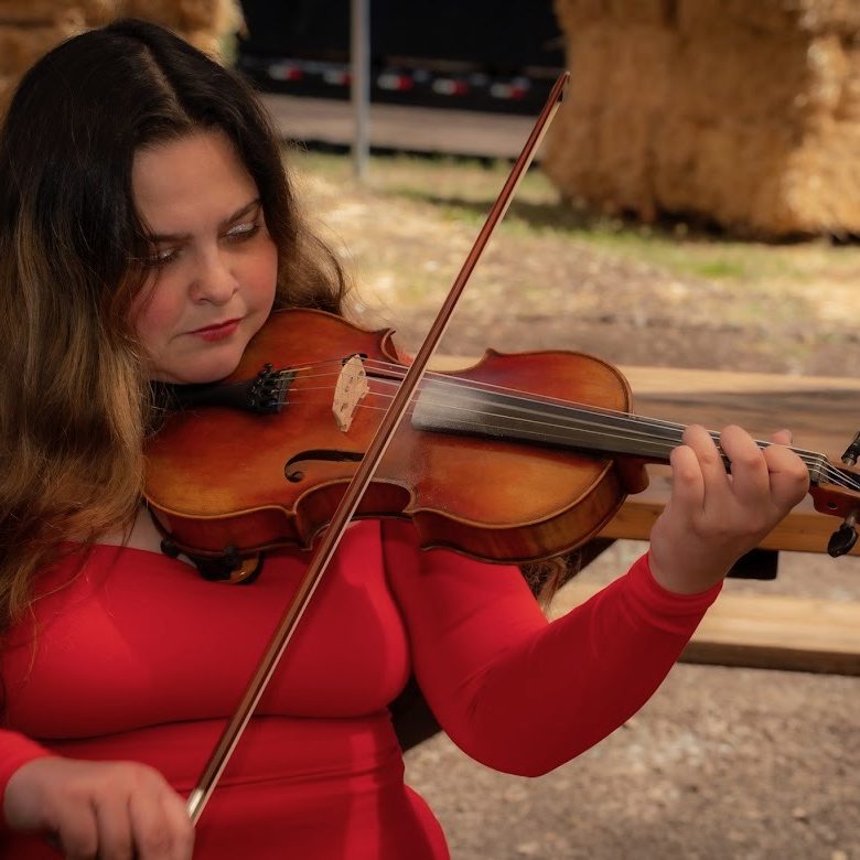 Welcome to High Key Wellness White woman wearing a long sleeved top. She has long, wavy brown hair with highlights and she is holding a violin/fiddle